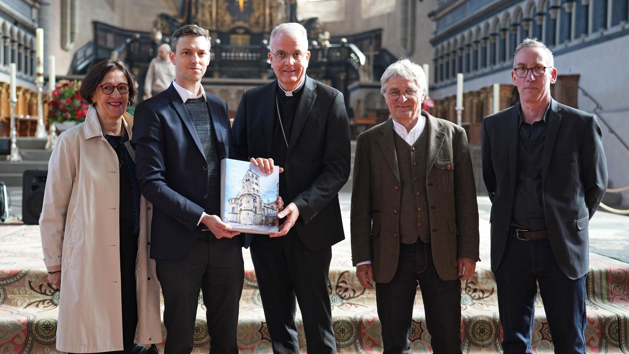 Vier Männer und eine Frau stehen im Trierer Dom vor dem Altar. Zwei Männer halten ein Buch in der Hand.